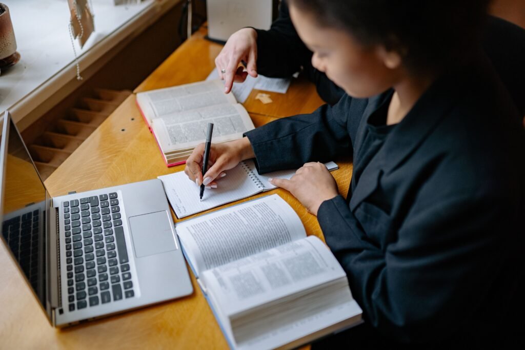 Two people reviewing open books and taking notes beside a laptop.