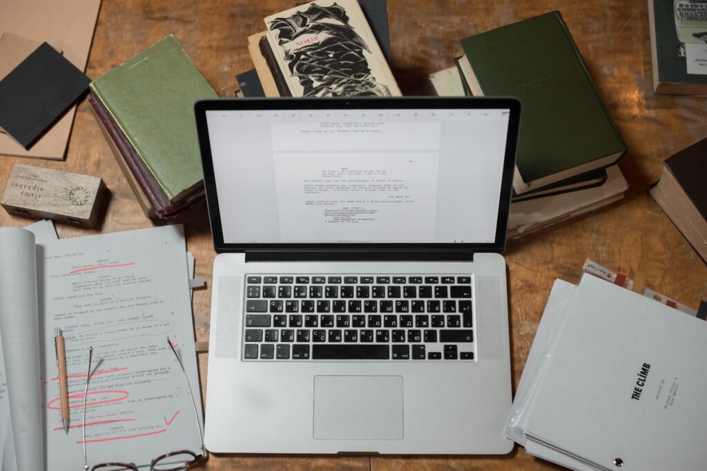 Laptop surrounded by books and manuscript documents on a wooden desk.